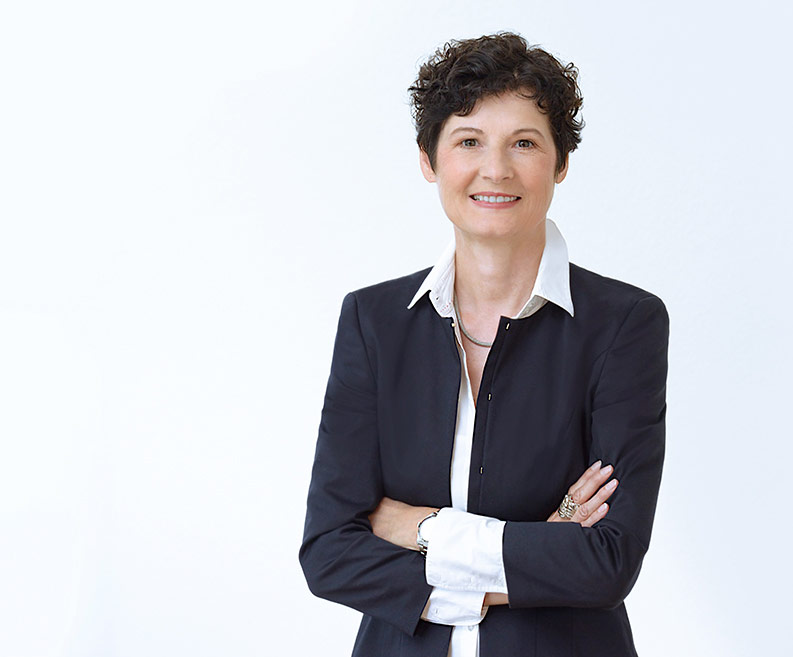 Portrait of DCZ Founder Member Dr. Carmen Zirngibl smiling with folded arms in dark suit jacket in front of light background.