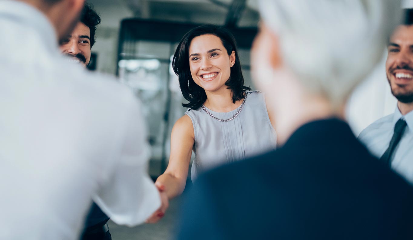 Scene of a greeting of a young woman in business attire surrounded by partly blurred people.